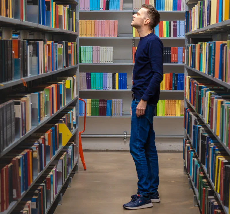 Person stood in a library looking at books, with filled bookshelves around them