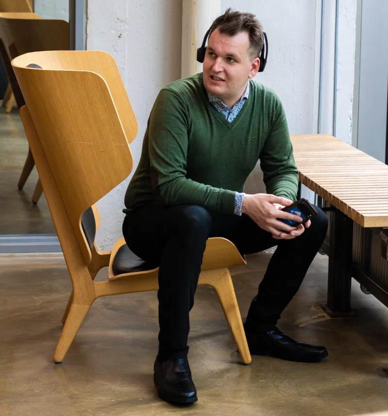 Picture of a person sat in a big chair, with a headset half on, and phone in hand, looking at someone or something off frame