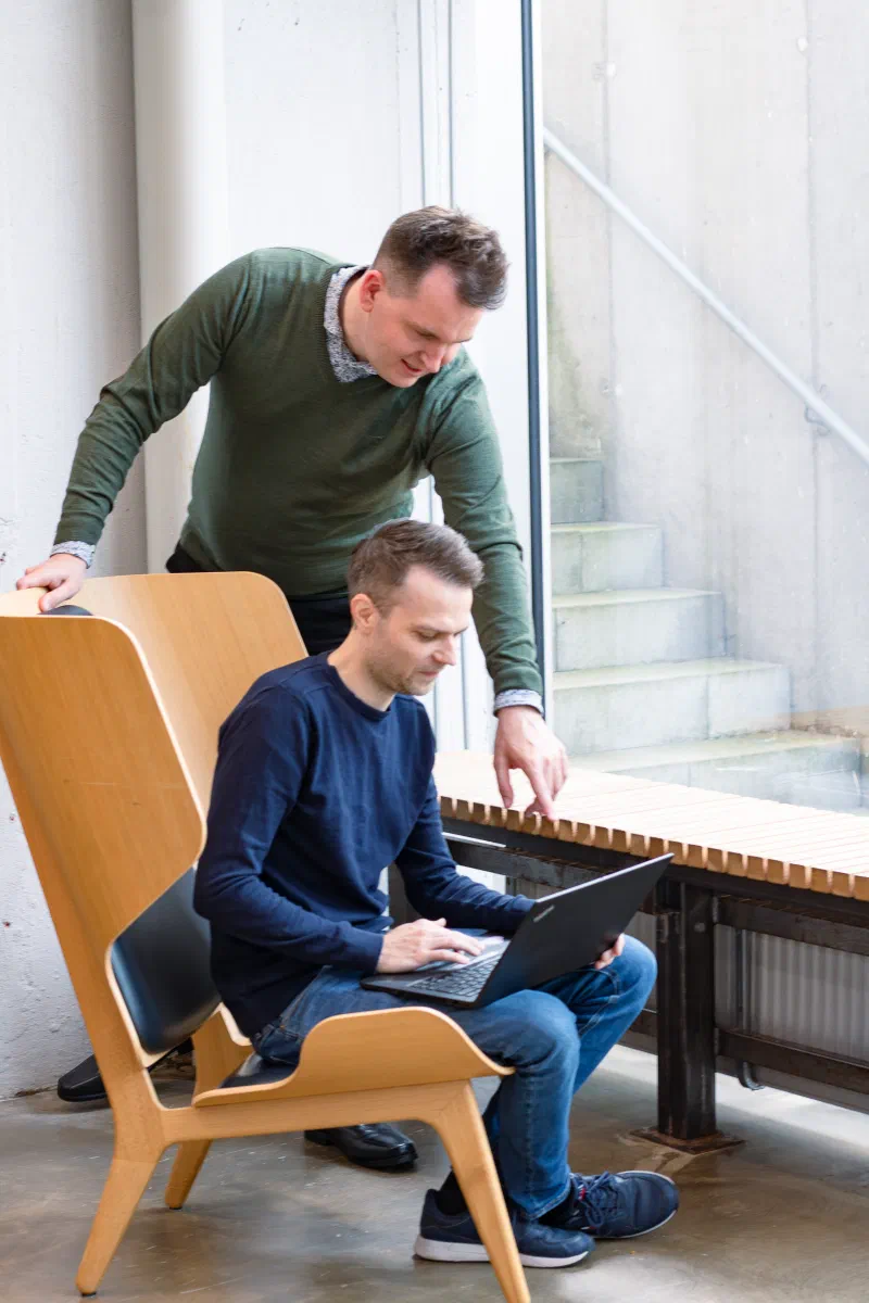 Picture of a person sat in a big chair with a laptop, and a guy looking over his shoulder, pointing at the screen