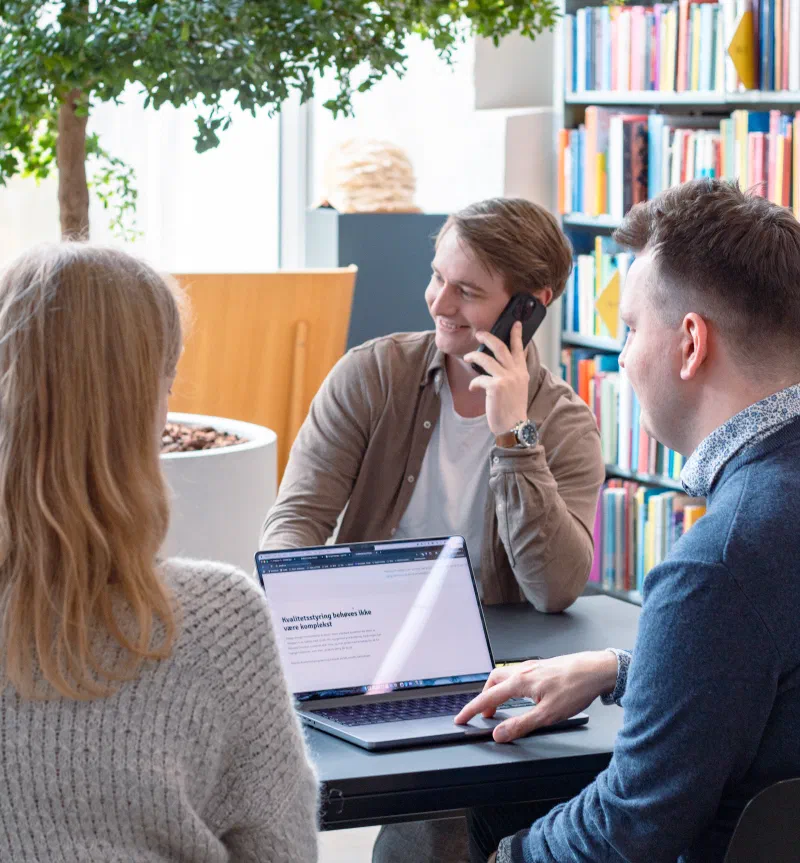 Picture of two people looking at a laptop at a table, and one person on the other side of the table on the phone