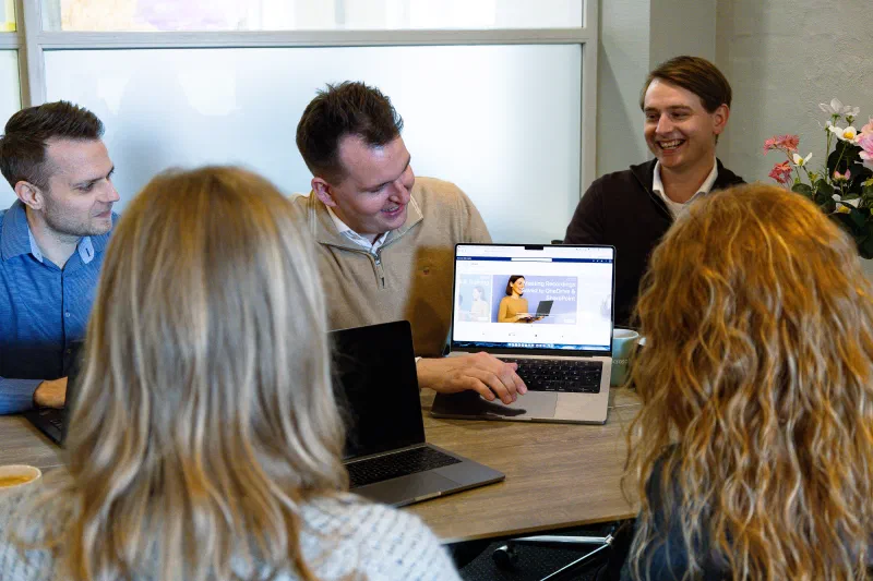 Picture of five people sat around a table, discussing something, three people on one side, and two on the other, the middle person on one side has his laptop flipped around, showing a SharePoint Online intranet